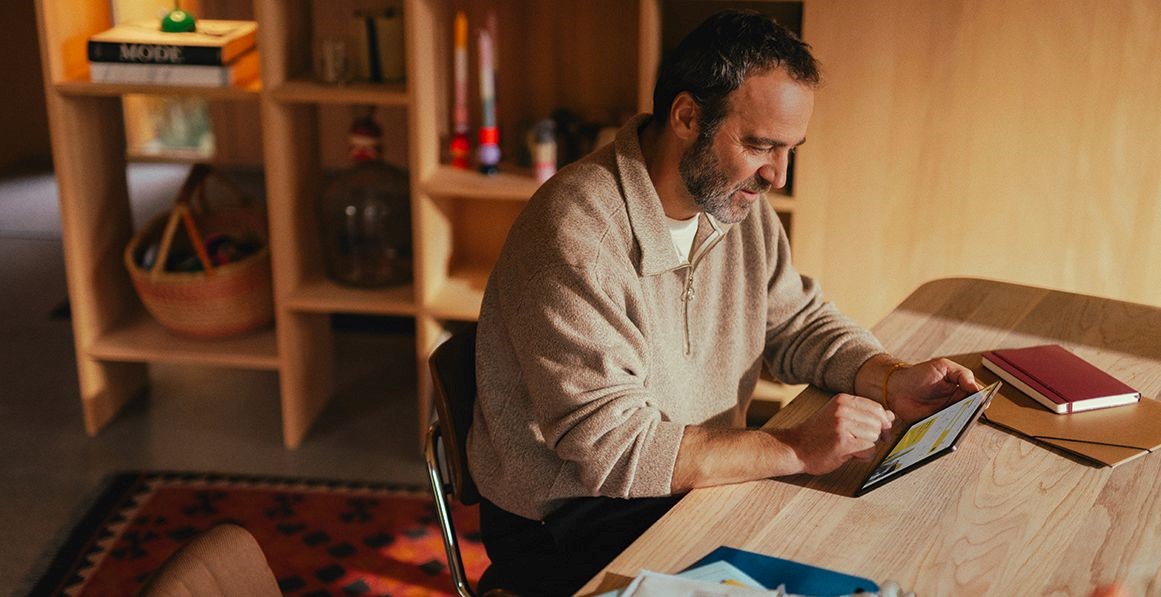 Ein Mann sitzt an einem Holztisch in einer wohnlichen Umgebung und nutzt die Post-App auf seinem Tablet, um Briefe digital zu empfangen.