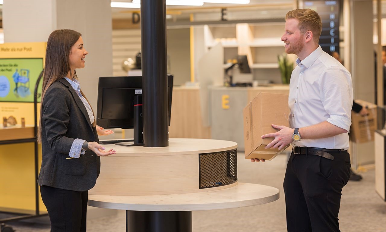Eine Post-Mitarbeiterin berät lächelnd einen Kunden, der ein Paket unter dem Arm hält, an einem modernen runden Schalter.