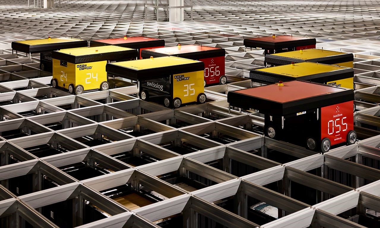 Mehrere kleine, rot-schwarze Logistik-Roboter bewegen sich auf einem silbernen Schienensystem in dem automatisierten Hochregallager in Villmergen.